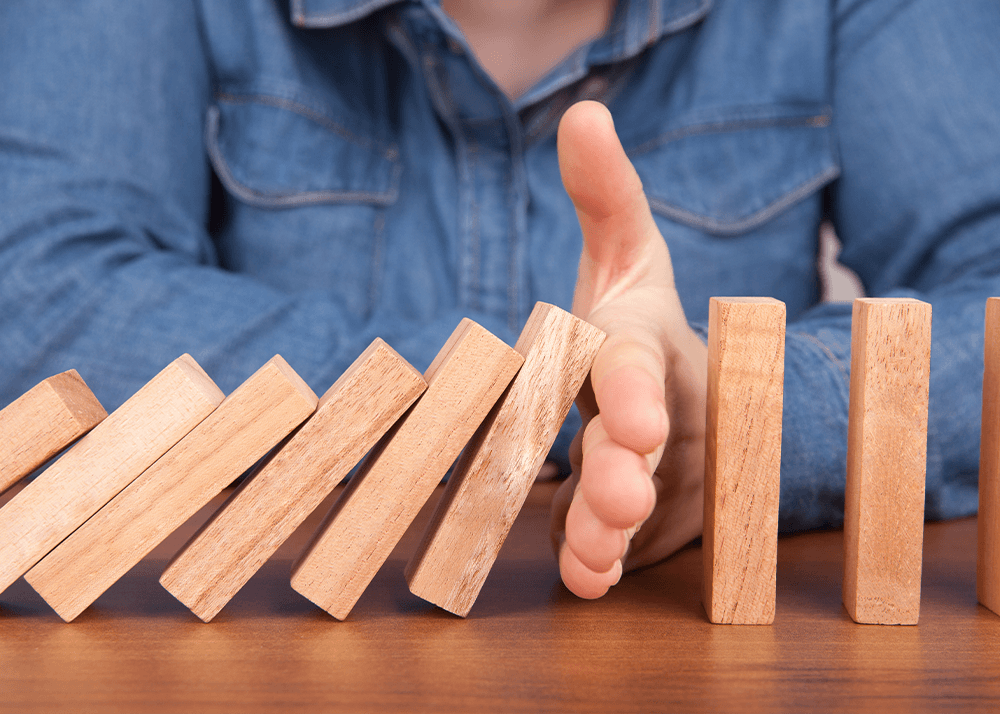 Reihe von Dominosteinen, bei denen eine Hand die Kette blockiert - eine einzige ausstehende Freigabe verzögert den gesamten Prozess