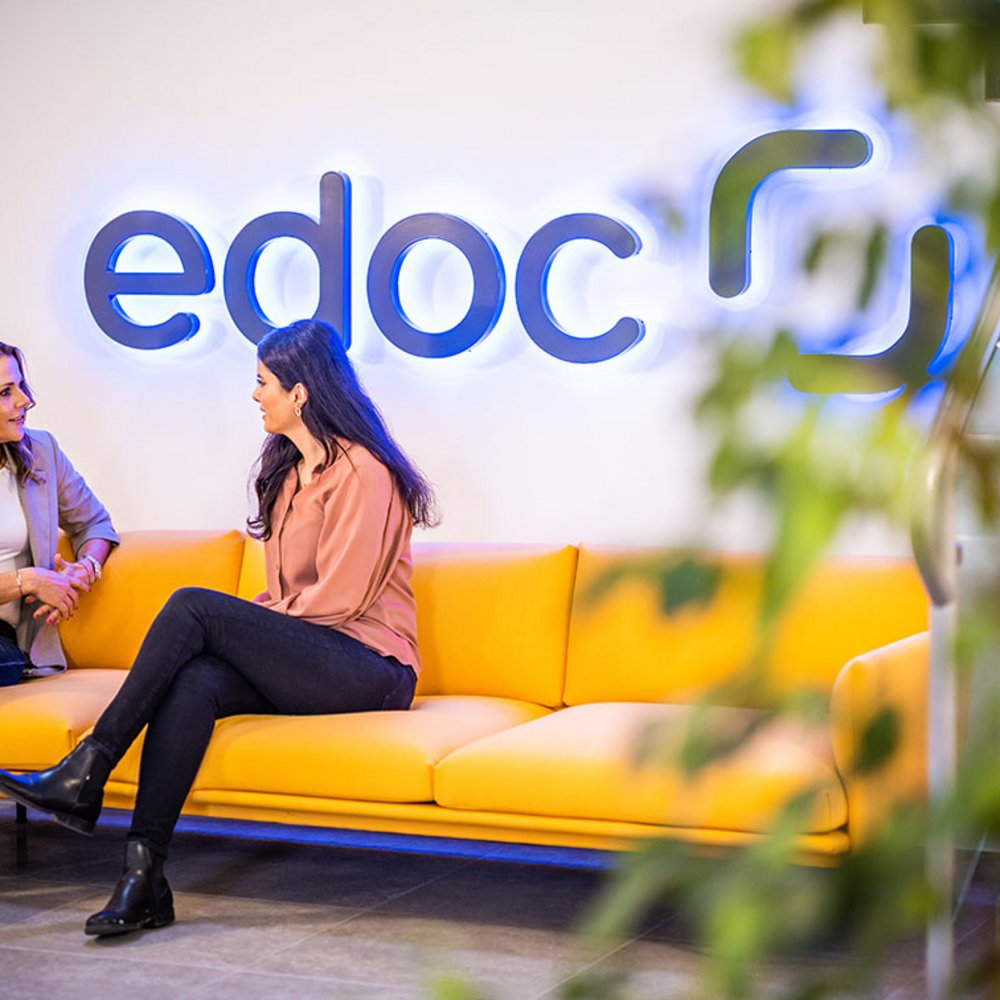 Zwei Mitarbeiterinnen von edoc sitzen auf einem gelben Sofa und unterhalten sich
